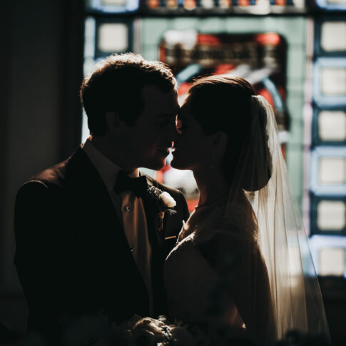 Sun shines through stained glass window of a church onto a bride and groom and their impending kiss.