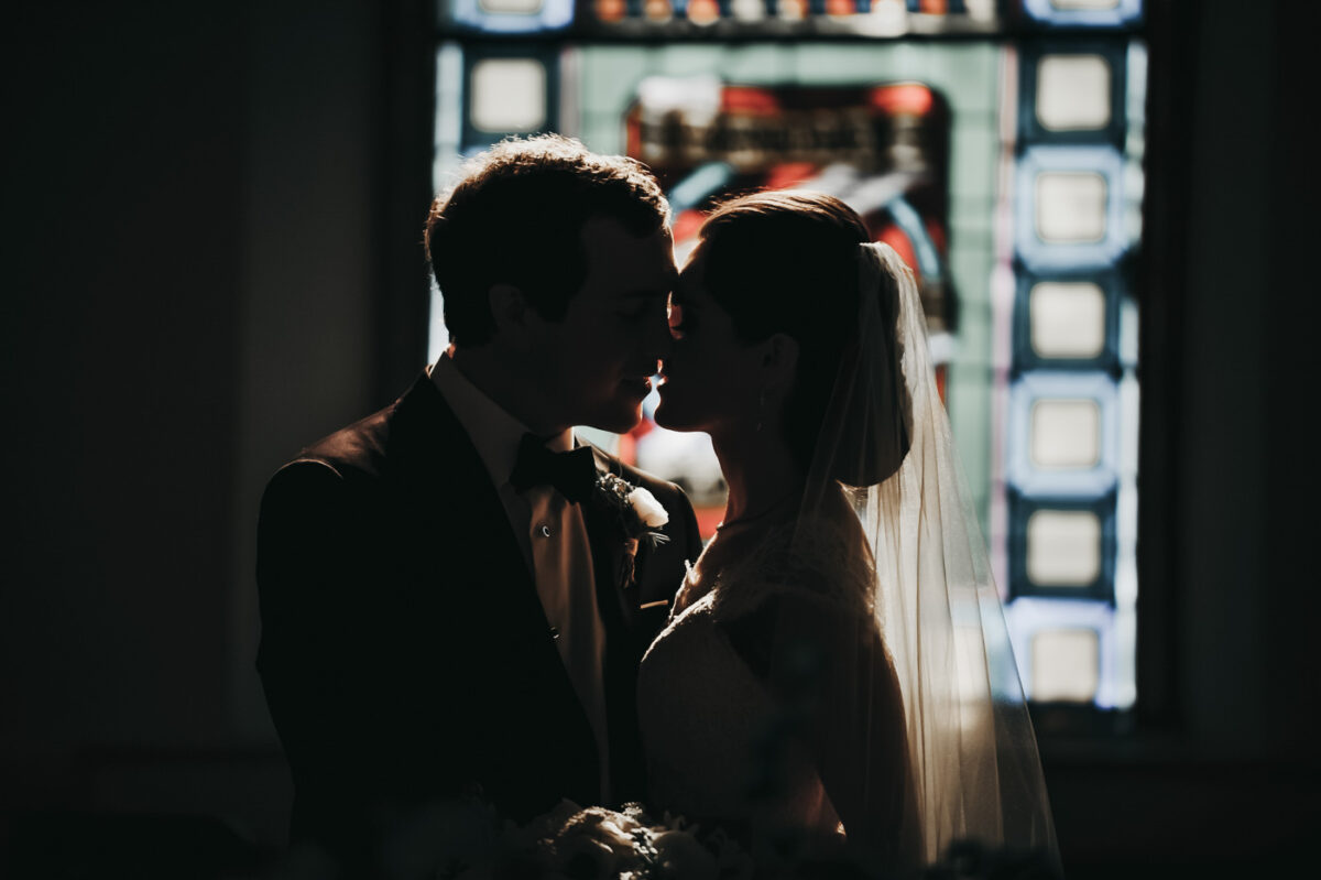 Sun shines through stained glass window of a church onto a bride and groom and their impending kiss.
