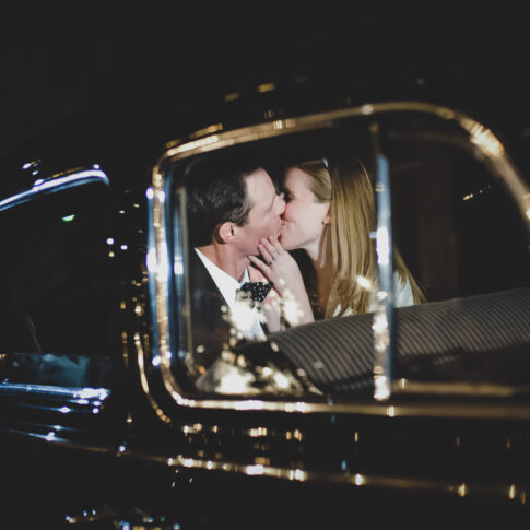 A bride and groom sneak a kiss in the back of their getaway car as they are leaving their wedding reception.