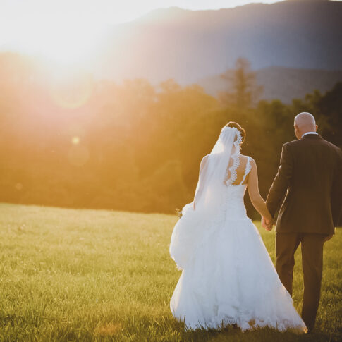 A bride and groom hold hands and walk away towards the setting sun over the NC mountains.