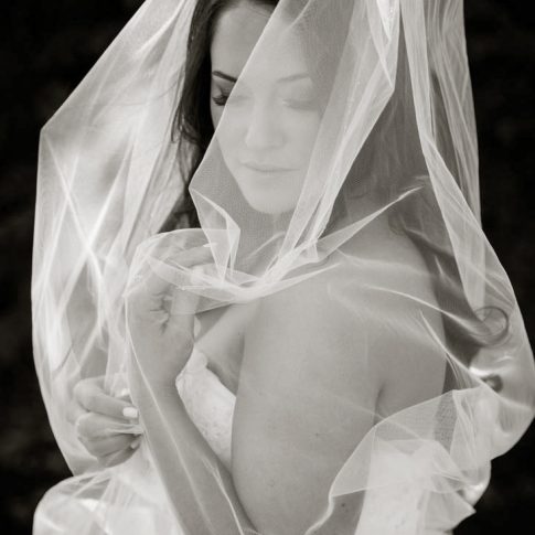 A bride, softly wrapped in her tulle veil, gazes down, showcasing her amazing bridal makeup, during a bridal portrait.