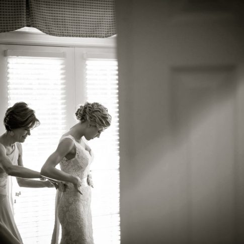 A mother carefully wraps a satin ribbon around her daughters waist and secures it in a bow in the back as her daughter prepares for her wedding day.