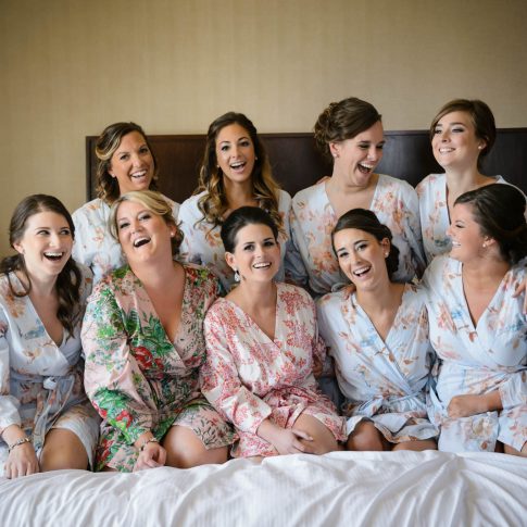 Bridesmaids join the bride in kneeling on the bed of the room they are getting ready for the wedding in and share a moment of laughter.