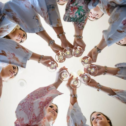 The bride and her bridesmaids share a toast while getting ready as this shot is taken from below, showcasing the bottoms of the glasses.