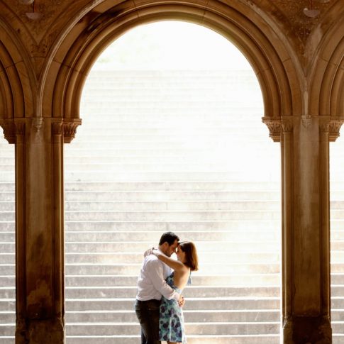 The soon-to-be-bride wraps her arms around her fiancé's neck underneath a magnificent archway during an engagement portrait session.