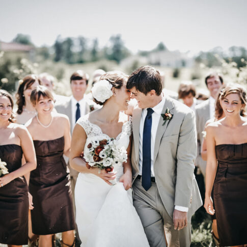 A bride and groom lean in for a kiss as their wedding party looks on in laughter while walking through the vineyards of Hotel Domestique.