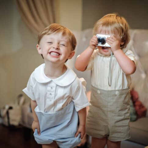 Two young boys at a wedding reception are filled with joy as one excitedly grins at the camera while another holds a small camera up to his eyes to take a photo.