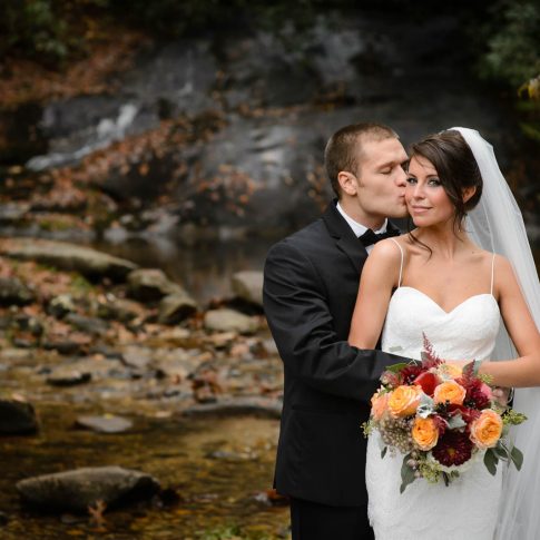 The bride softly glances at the camera as her groom kisses her cheek while they stand embraced in front of a waterfall in Lonesome Valley in Cashiers, NC.