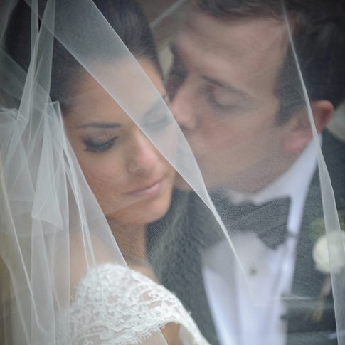 A bride and groom snuggle under her veil as the groom kisses the bride's cheek.