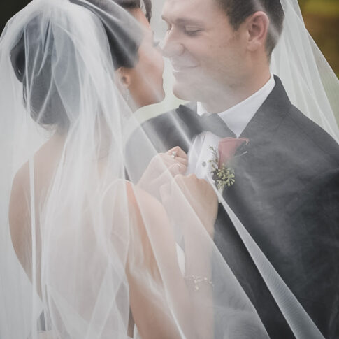 The bride and groom happily smile at each other as they stand draped by the brides veil.