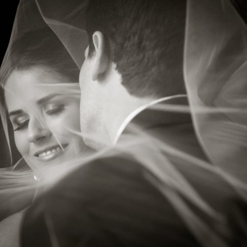 A groom kisses his bride on the cheek as they are both surrounded by her tulle veil.