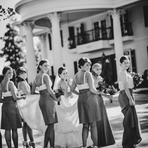 Bridesmaids carefully lift the brides train as they turn back and smile outside of the wedding venue, the Ryan Nicholas Inn.