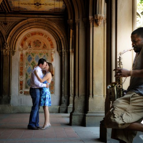 Underneath beautiful historic arches in NYC with a saxophone player in the foreground, an engaged couple shares a romantic kiss.