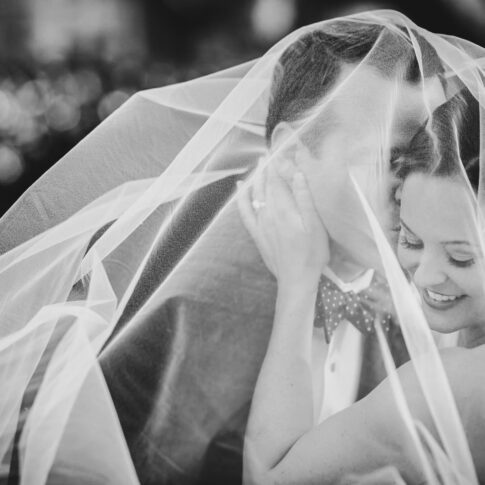 A groom plants a quick kiss on the cheek of his wife as they are both covered in the tulle of her veil.