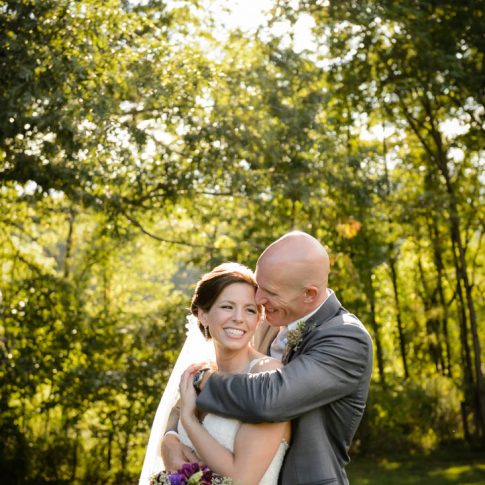 A groom hugs his new wife from behind as they smile and stand surrounded by lush greenery.