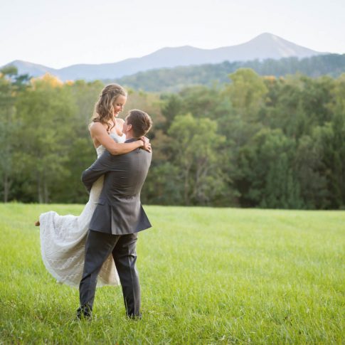 A groom swoops his bride up in his arms as they stand in a lush field in Candler, NC.