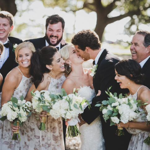 Husband and wife hug and kiss while being surrounded by their wedding party laughing.