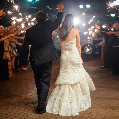 A bride and groom jovially wave goodbye to their guests who wave sparklers in celebration of sending off the happy couple.