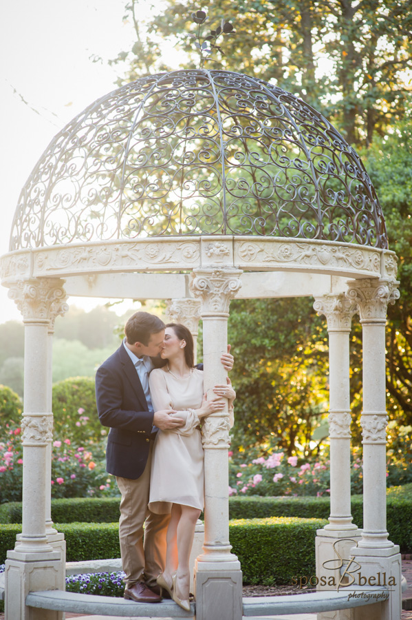 Engaged couple shares a kiss underneath a wrought iron pavilion in the scenic Furman Rose Garden. 