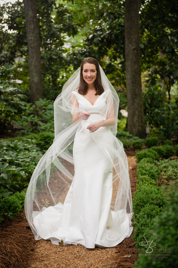 Bride carefully wraps her veil around herself while standing in a garden. 