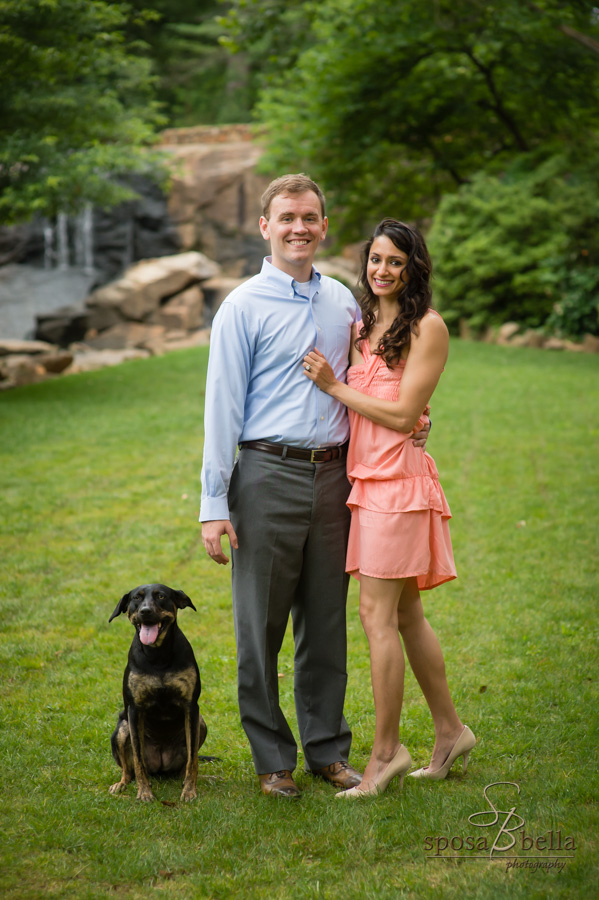 Engaged couple stands and smile with their dog sitting beside them.