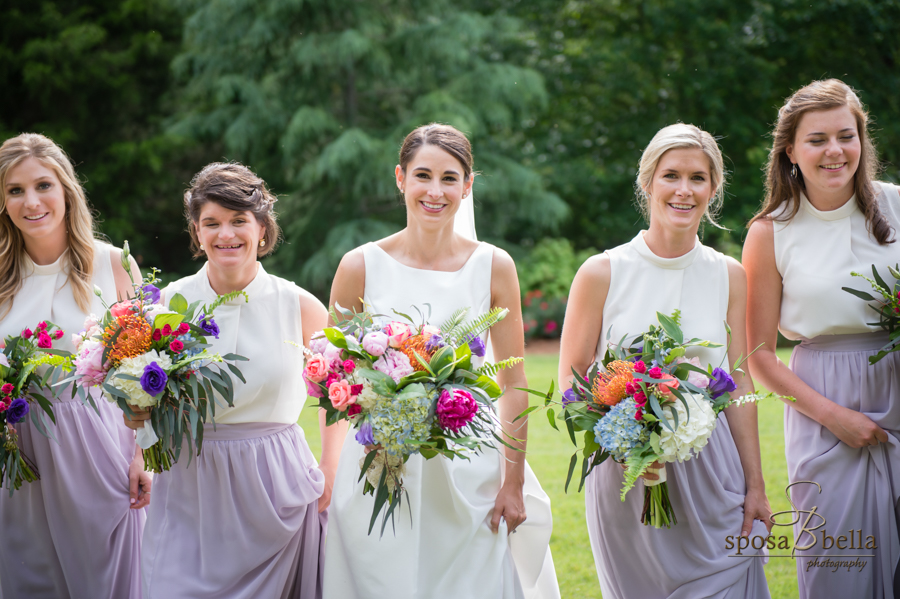 The bride and her bridesmaids all smile as they walk along the grass. 